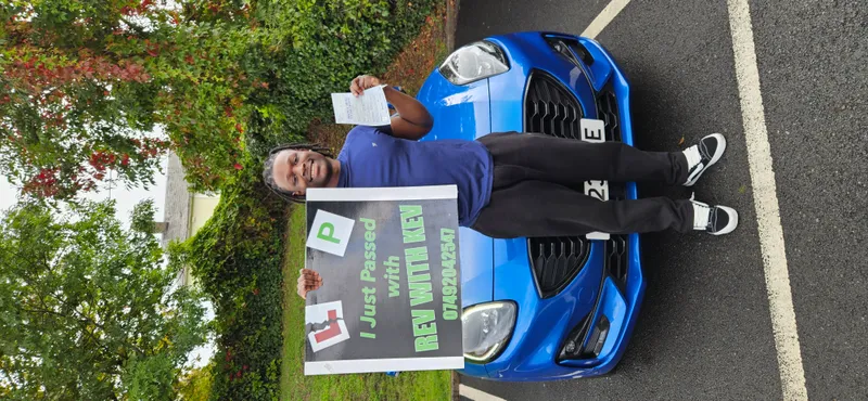 Happy student holding I Just Passed sign next to blue Rev with Kev driving school car in Aberdare